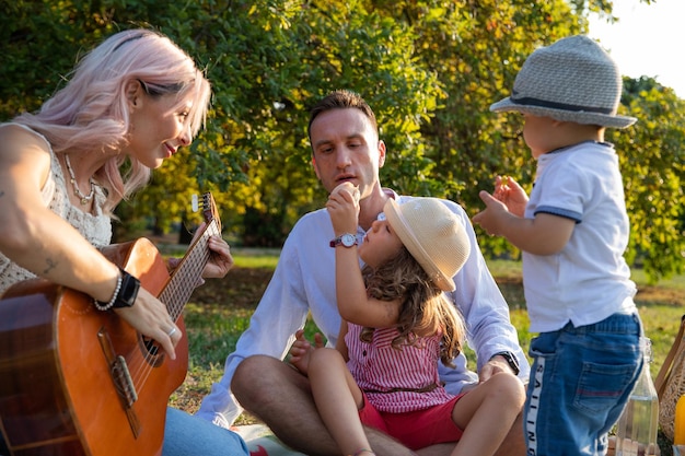 Famille de quatre personnes avec de jeunes enfants dans le parc en train de pique-niquer pendant que maman joue de la guitare