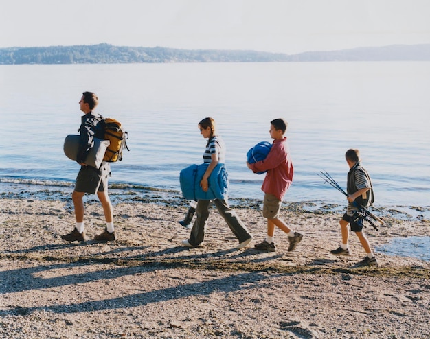 Photo famille de quatre personnes transportant du matériel de camping marchant sur la plage au crépuscule