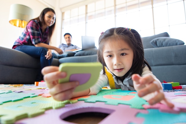 Famille de race mixte restant ensemble, père caucasien travaillant à la maison sur un canapé tandis que petite jolie fille mignonne jouant au jeu d'alphabet éducatif sur le sol et mère asiatique regardant avec amour. Idée pour une famille chaleureuse