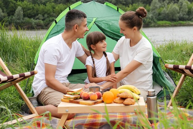 Famille se reposant au bord de la rivière Camping heureux près de la rivière dans le parc naturel Couple avec fille assise près de la tente Week-ends en plein air