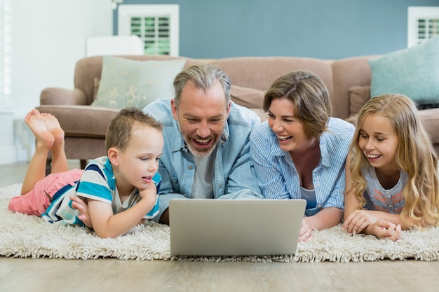 Famille souriante à l'aide d'un ordinateur portable en position couchée sur un tapis dans le salon