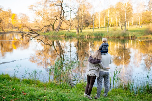 Famille de style de vie par derrière avec leur enfant sur les épaules du père debout au bord du lac