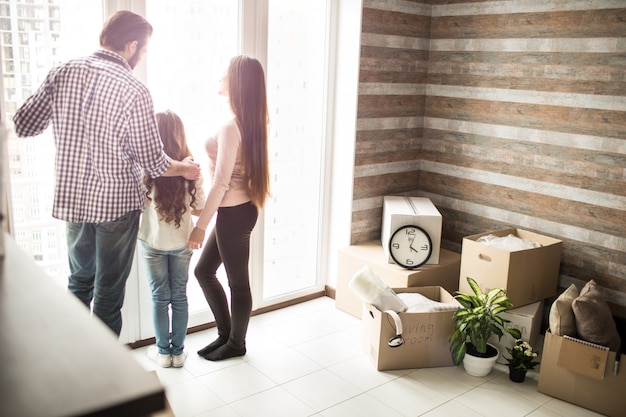 Une Famille Sympathique Se Tient Près D'une Fenêtre Et Regarde à L'extérieur. Il Y A Des Boîtes D'homme Sur Le Côté Droit De L'appartement. Les Gens Parlent De Quelque Chose.