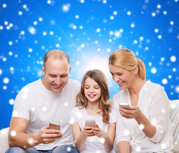 famille, vacances de noël, technologie et personnes - mère souriante, père et petite fille avec des smartphones sur fond bleu neigeux