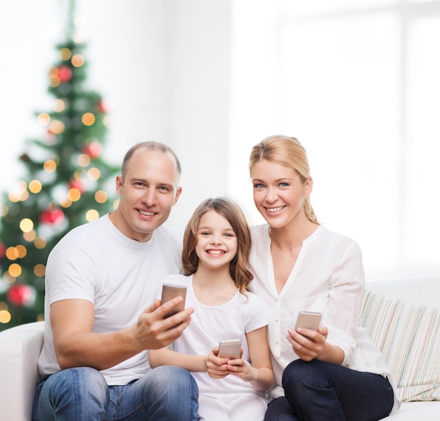 famille, vacances, technologie et personnes - mère, père et petite fille souriants avec des smartphones sur fond de salon et d'arbre de noël