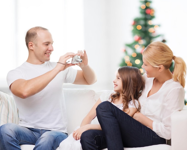 famille, vacances, technologie et personnes - mère souriante, père et petite fille avec caméra sur fond de salon et arbre de noël