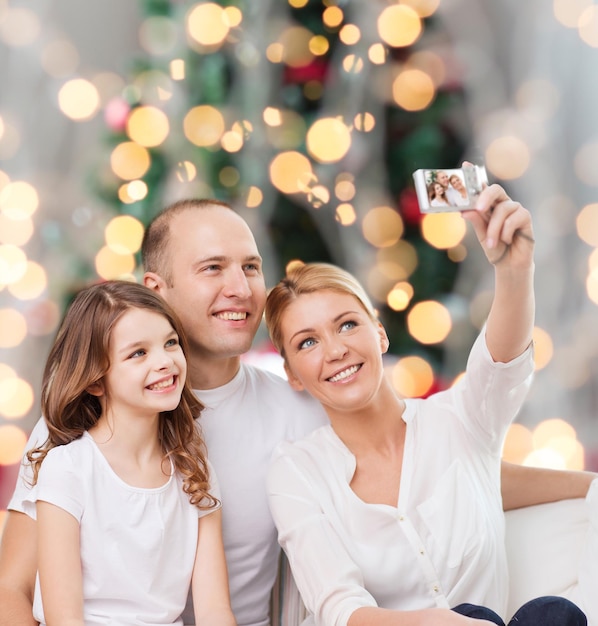 famille, vacances, technologie et personnes - mère souriante, père et petite fille faisant selfie avec caméra sur fond de lumières de sapin de noël