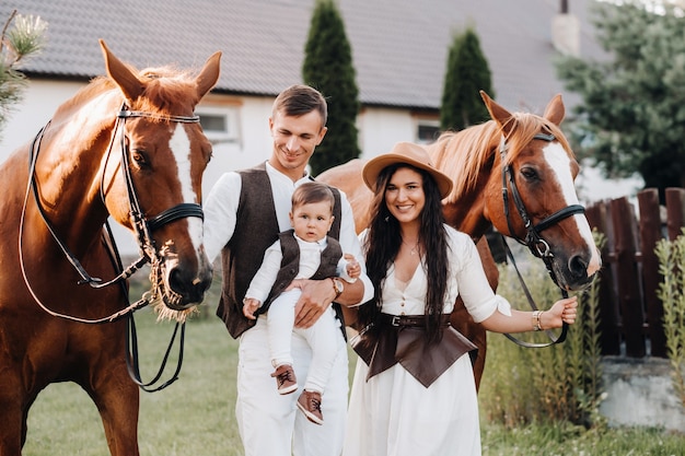 Une famille en vêtements blancs avec leur fils se tient près de deux beaux chevaux dans la nature. Un couple élégant avec un enfant est photographié avec des chevaux.
