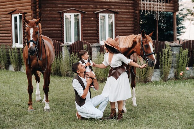 Une famille en vêtements blancs avec leur fils se tient près de deux beaux chevaux dans la nature. Un couple élégant avec un enfant est photographié avec des chevaux