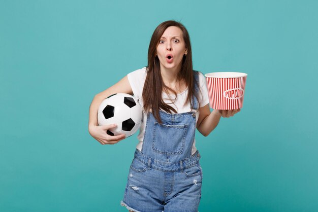 Fan de football choqué de jeune fille regardant l'équipe favorite de soutien de match avec ballon de football, seau de pop-corn isolé sur fond bleu turquoise. Émotions des gens, concept de mode de vie de loisirs familiaux sportifs.