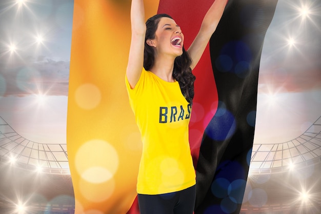 Fan de football excité en t-shirt brésilien tenant le drapeau allemand contre un grand stade de football sous un ciel bleu nuageux