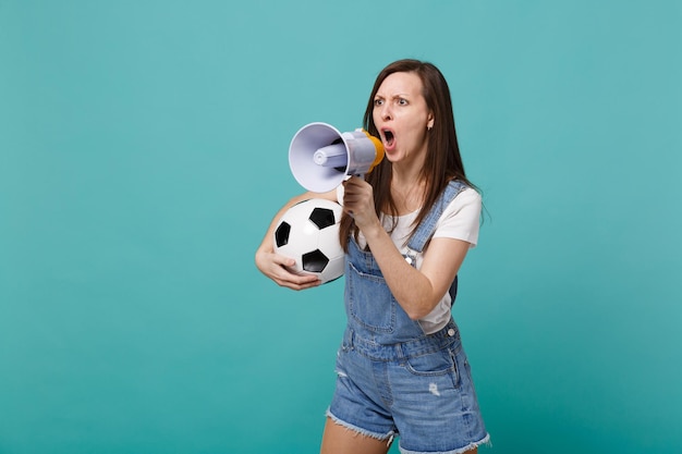 Fan de football femme en colère crier dans un mégaphone, soutenir l'équipe préférée avec un ballon de football isolé sur fond bleu turquoise. Émotions des gens, concept de mode de vie de loisirs familiaux sportifs. Maquette de l'espace de copie.