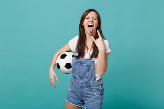 Fan de football folle jeune femme égayer l'équipe préférée de soutien avec ballon de football montrant la langue, cornes en geste isolé sur fond bleu turquoise. Émotions des gens, concept de loisirs sportifs en famille.