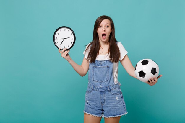 Fan de football de jeune femme abasourdie tenant un ballon de football, horloge ronde isolée sur fond de mur bleu turquoise en studio. Le temps presse. Émotions des gens, concept de mode de vie de loisirs familiaux sportifs.