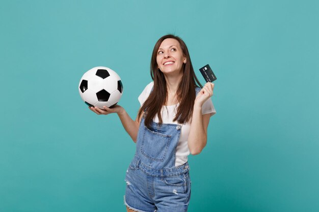 Un fan de football de jeune femme réfléchie soutient l'équipe préférée avec un ballon de football, une carte bancaire de crédit, levant isolé sur fond bleu turquoise. Émotions des gens, concept de mode de vie de loisirs familiaux sportifs.