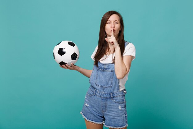 Un fan de football de jolie femme soutient l'équipe préférée avec un ballon de football et dit tais-toi, tais-toi avec le doigt sur les lèvres chut geste isolé sur fond bleu turquoise. Les émotions des gens sport concept de loisirs en famille.