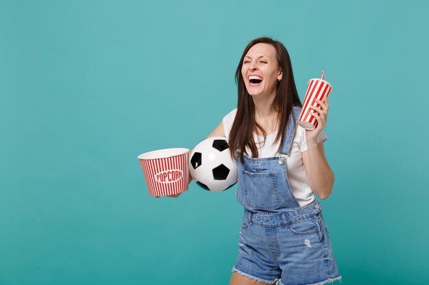 Une fan de football qui rit en regardant un match, soutient son équipe préférée avec un ballon de football, un seau de pop-corn, une tasse de cola ou de soda isolé sur fond bleu turquoise. Concept de mode de vie sport loisirs familiaux.