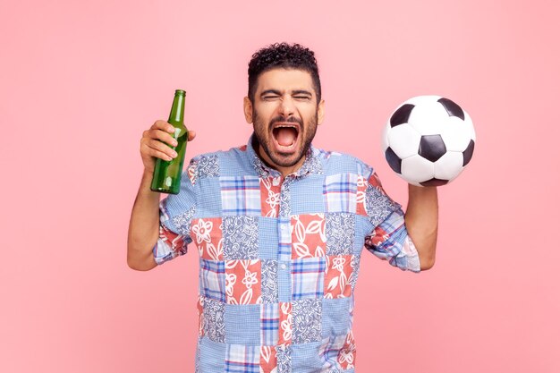 Un fan fou portant une chemise bleue de style décontracté criant joyeusement, célébrant la victoire de son équipe préférée, tenant dans les mains une balle et une bouteille d'alcool. Tourné en studio intérieur isolé sur fond rose.