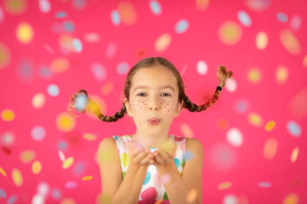 Fantaisie enfant avec des nattes soufflant des confettis contre le mur rose.