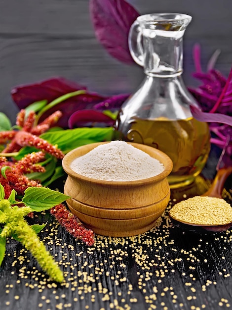 Farine d'amarante dans un bol, graines dans une cuillère et huile dans une carafe, feuilles et fleurs brunes, vertes et violettes de plante sur fond de planche de bois