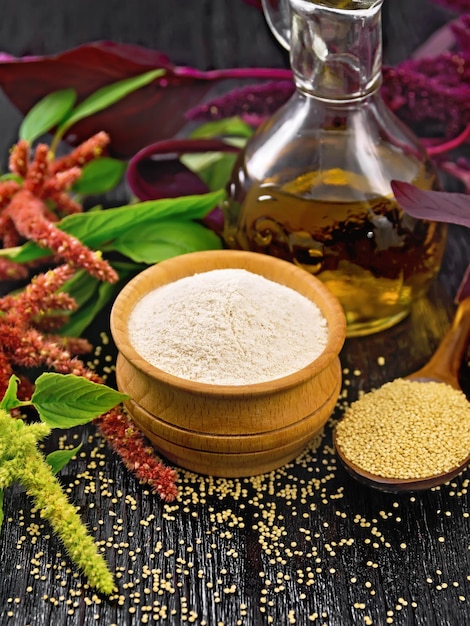 Farine d'amarante dans un bol graines dans une cuillère et huile dans une carafe fleurs marron vert et violet de plante sur fond de planche de bois