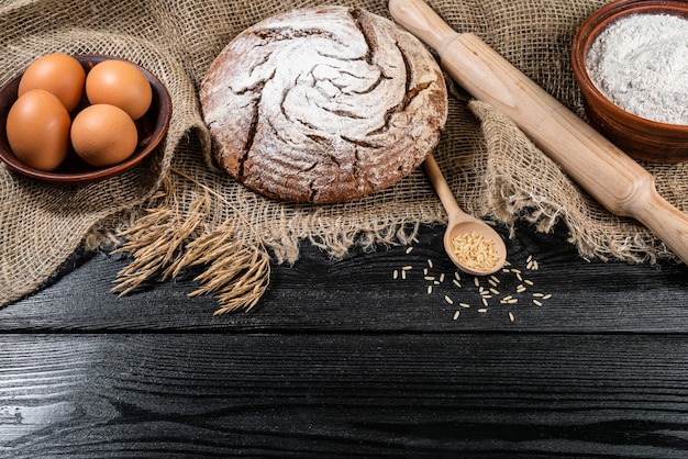 Farine dans un bol en bois sur une table en bois foncé avec des épillets de blé, des œufs et du lait, vue de dessus avec copie espace