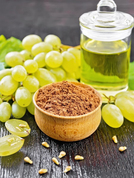 Farine de pépins de raisin dans un bol, huile dans un bocal et baies de raisins sur fond de planche de bois noir