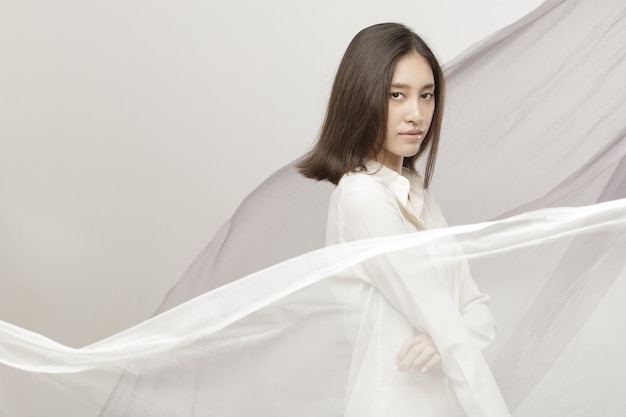 Fashion Beauty Woman a les cheveux noirs courts et raides et exprime des sentiments. Portrait d'une fille thaïlandaise asiatique portant une chemise blanche avec une vague de tissu sur le vent, un jet de tissu flottant dans le ciel sur fond blanc cassé