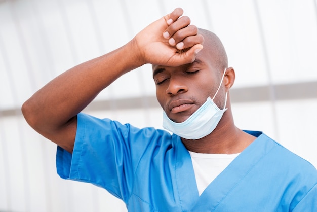 Fatigué après une longue opération. Jeune médecin africain fatigué en uniforme bleu touchant son front avec la main et gardant les yeux fermés