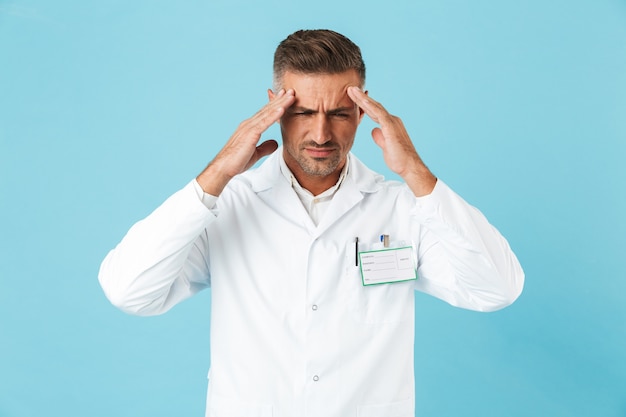 Fatigué de l'homme médecin en uniforme debout isolé sur mur bleu, souffrant d'un mal de tête