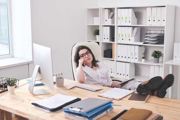 Fatigué de jeune femme assise dans une pose détendue à table et regardant l'écran de l'ordinateur