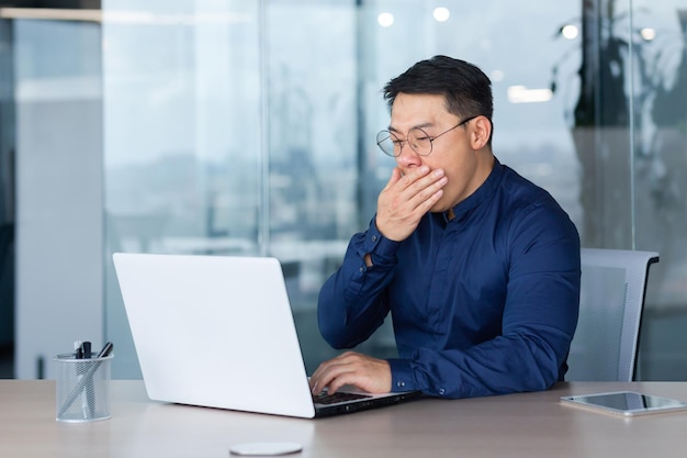 Fatigué jeune homme d'affaires asiatique bâille sur le lieu de travail, il est assis endormi au bureau dans le bureau