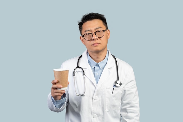 Fatigué malheureux médecin chinois d'âge moyen en blouse blanche et verres avec une tasse de café à emporter