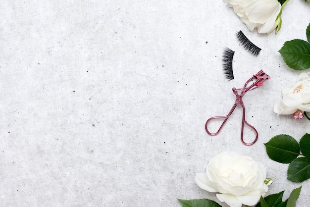 Faux cils avec pince à épiler rose sur fond gris avec fleurs blanches, vue de dessus. Notion de beauté. espace de copie