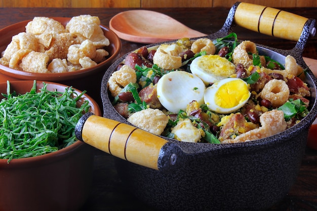 Feijao Tropeiro, plat typique de la cuisine brésilienne, à base de haricots, bacon, saucisses, chou vert, œufs, sur une table en bois rustique.