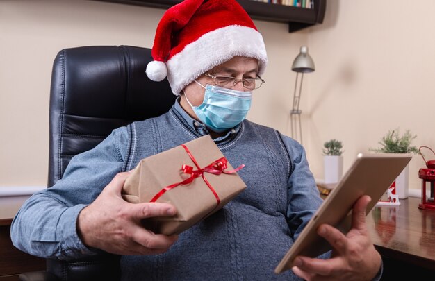 Félicitations de Noël en ligne. Man in santa claus hat parle à l'aide de la tablette pour les amis et les enfants des appels vidéo. La salle est décorée de façon festive. Noël pendant le coronavirus.