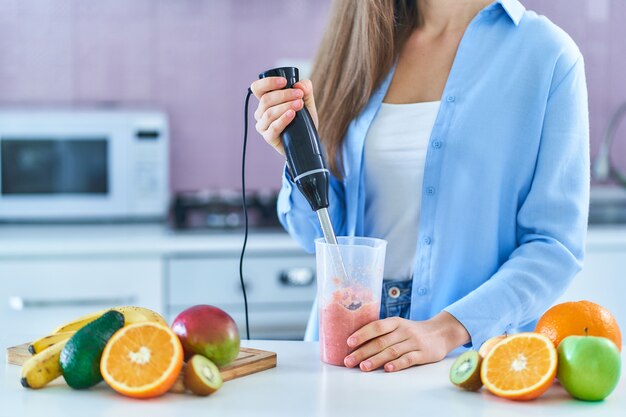 La femelle utilise un mélangeur à main pour mélanger les fruits frais pour préparer un smoothie diététique dans la cuisine à la maison