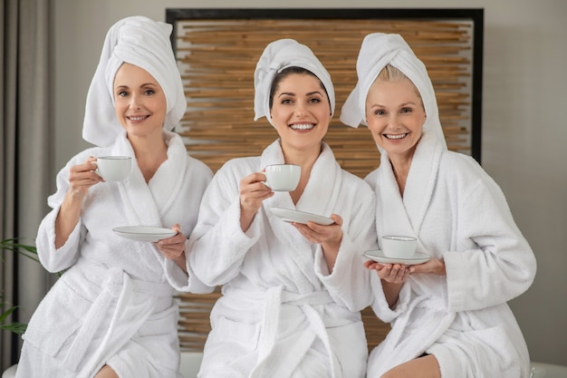 La féminité. Belles femmes joyeuses adultes en peignoirs et serviettes sur la tête avec des tasses assises se reposant en passant du temps au spa