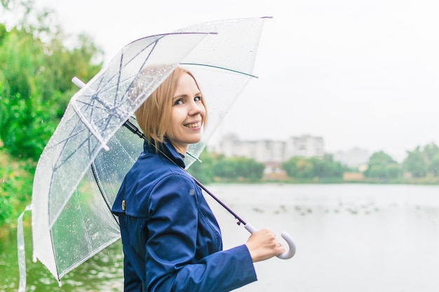 Une femme de 3040 ans sourit dans un imperméable bleu sous un parapluie transparent à l'automne près du lac