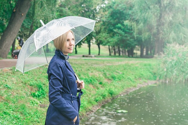 Une femme de 3040 ans sourit dans un imperméable bleu sous un parapluie transparent à l'automne près du lac