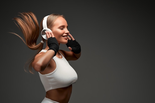 Femme avec des abdominaux parfaits portant des écouteurs et des vêtements blancs debout dans la salle de sport isolé sur fond gris