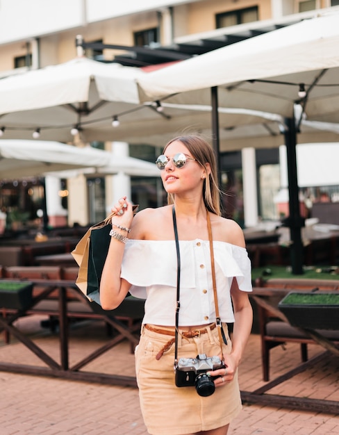 Femme accro du shopping à la mode dans des vêtements élégants avec appareil photo rétro tenant des sacs à provisions sur la rue de la ville