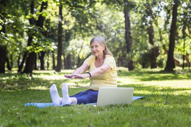 Femme active aux cheveux gris dans le parc faisant du fitness assis sur le tapis et l'herbe à l'aide d'un ordinateur portable pour des cours en ligne dans le parc