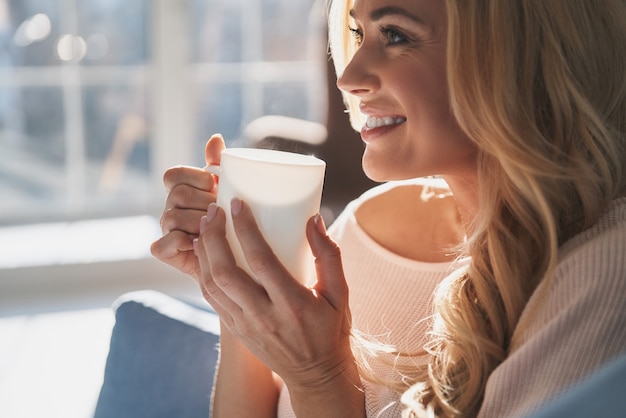 Femme adorable. Jolie jeune femme tenant une tasse et regardant loin avec le sourire alors qu'elle était assise sur le canapé à la maison