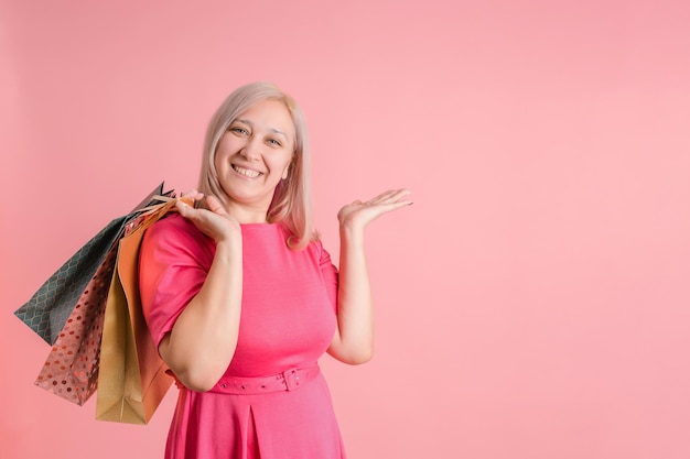 Une femme adulte de 40 ans avec des sacs à provisions et une paume ouverte montre sur fond rose