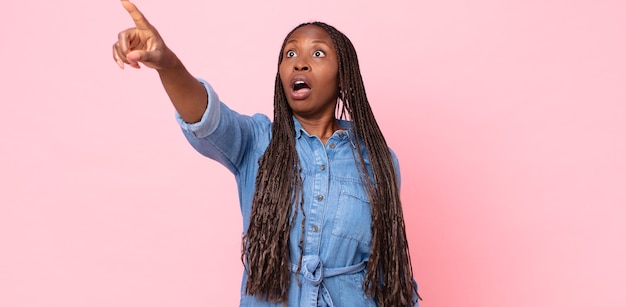 Femme adulte afro noire se sentant choquée et surprise, pointant du doigt et regardant vers le haut avec admiration avec un regard étonné et bouche bée