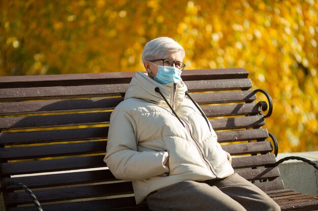 Femme adulte âgée assis sur un banc de parc à l'automne portant un masque médical