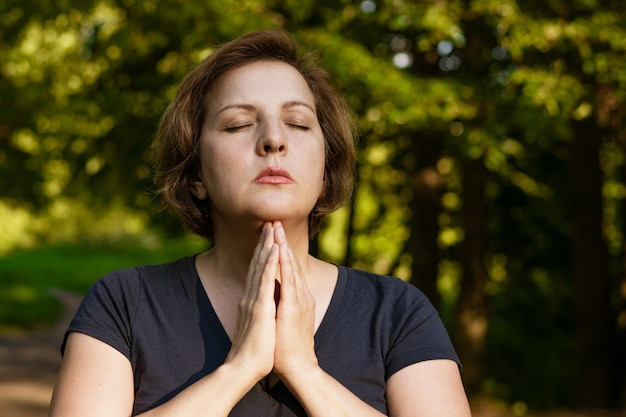 Femme adulte aux cheveux courts médite avec les paumes pliées dans le parc avec les yeux fermés dans les rayons du soleil gaggle en été