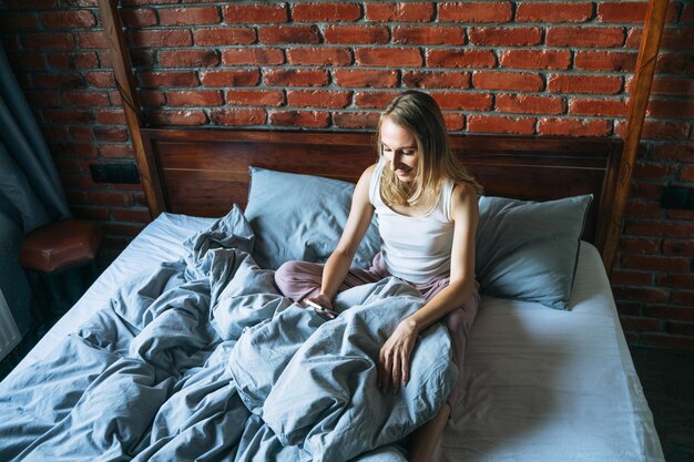 Une femme adulte aux cheveux longs blonds en pyjama utilisant un téléphone portable assise sur le lit dans une chambre au grenier à la maison