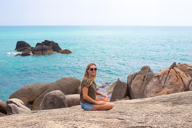 Une femme adulte aux cheveux longs est assise sur une pierre au bord de la mer et sourit Voyages et tourisme